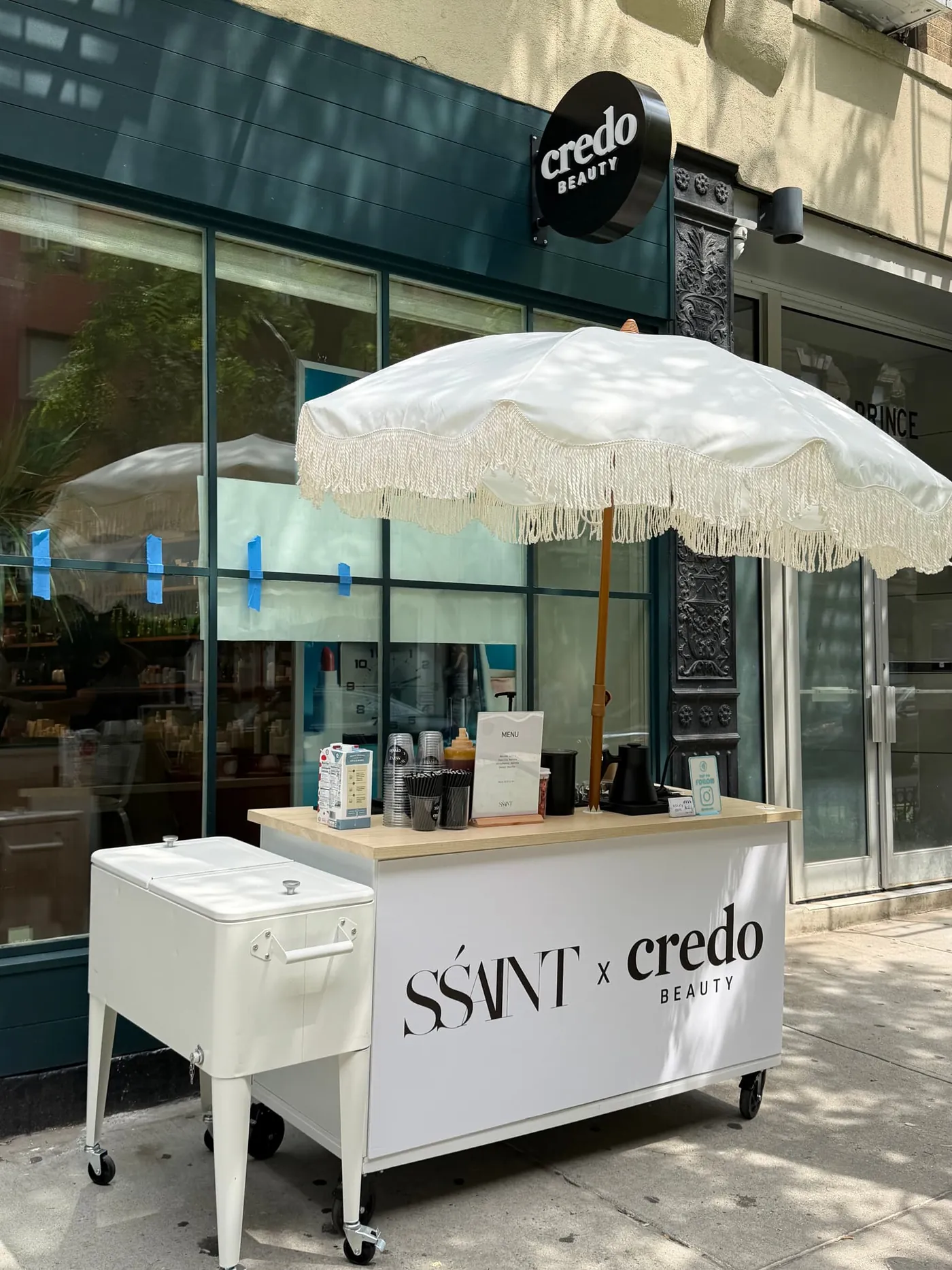 Melu's Cafe cart with a white fringed umbrella, branded SSAINT x Credo Beauty, outside a NYC storefront