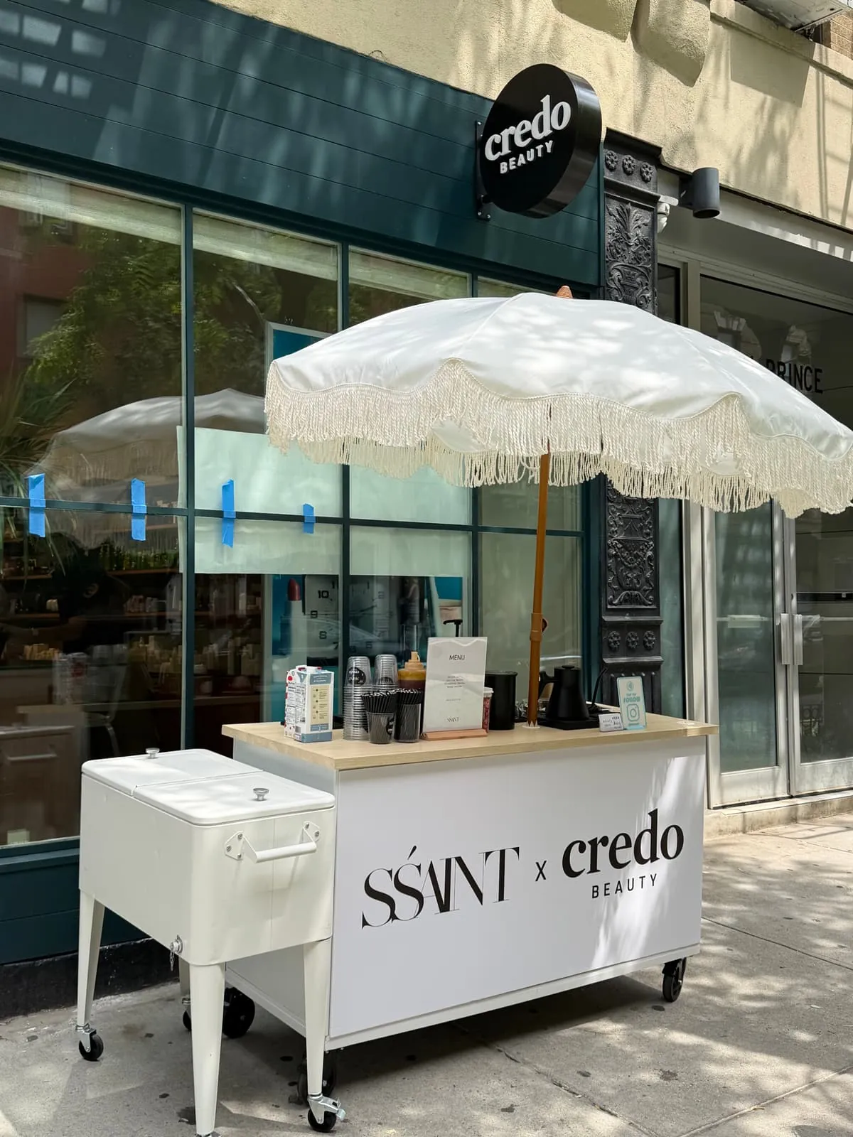 Coffee and matcha cart under a fringed umbrella on a NYC sidewalk outside Credo Beauty